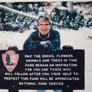 Man in jacket by park sign in snow.