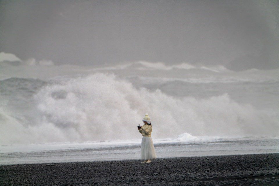 Woman in white stands on black sand beach.